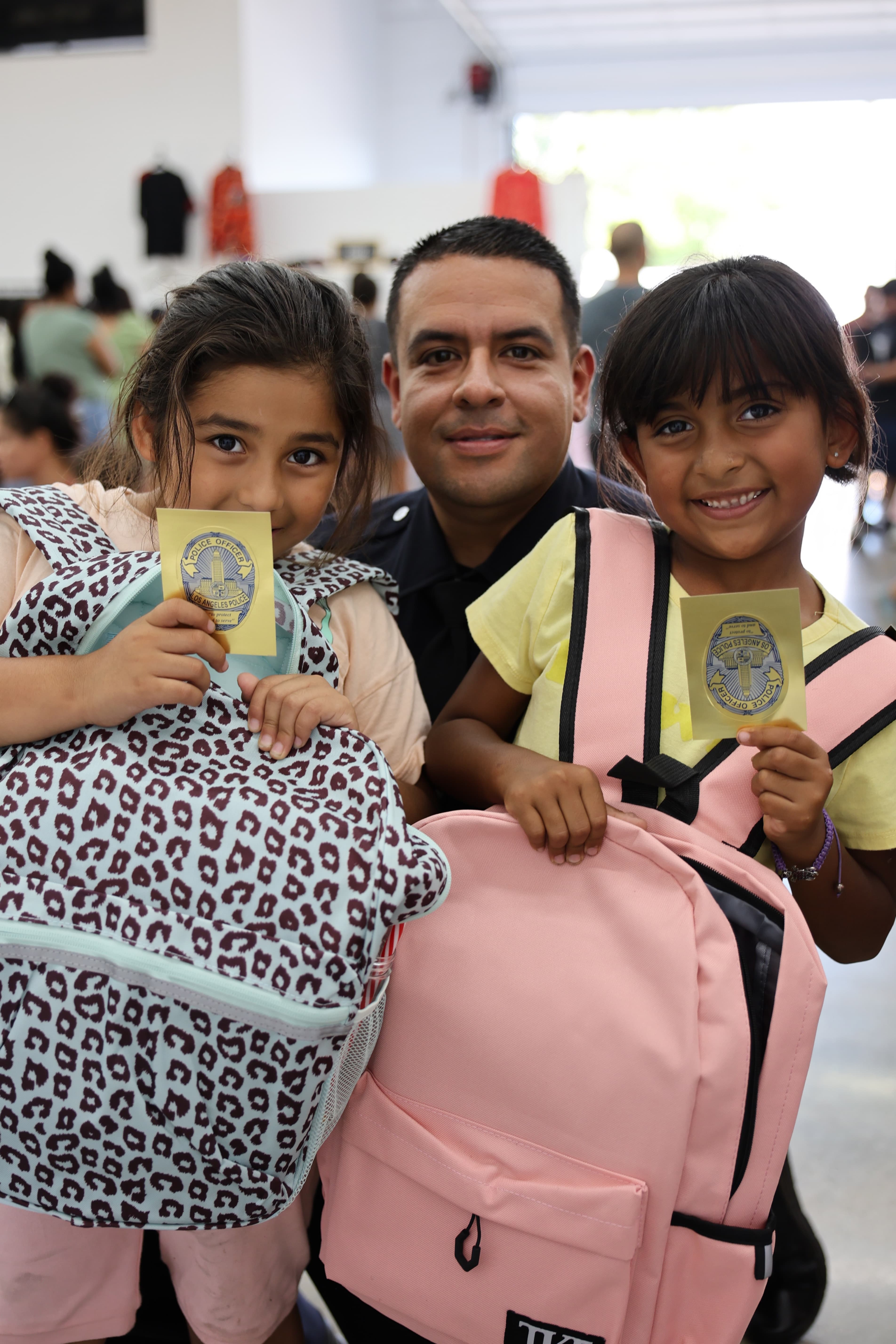 Two smiling children holding new backpacks and LAPD junior-officer badges beside a Los Angeles police officer at a Trina's Kids Back-to-School Drive