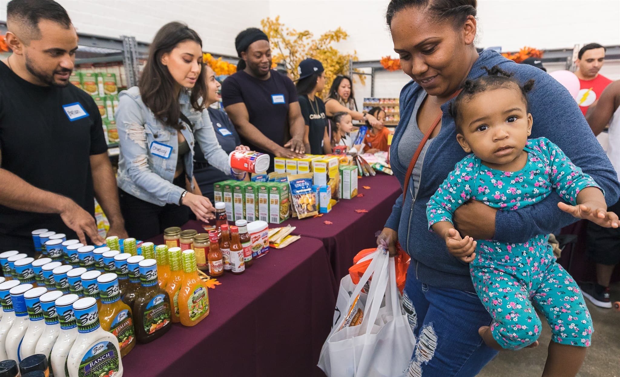 A parent carrying a toddler past tables of groceries and pantry staples at a Trina's Kids Thanksgiving food drive